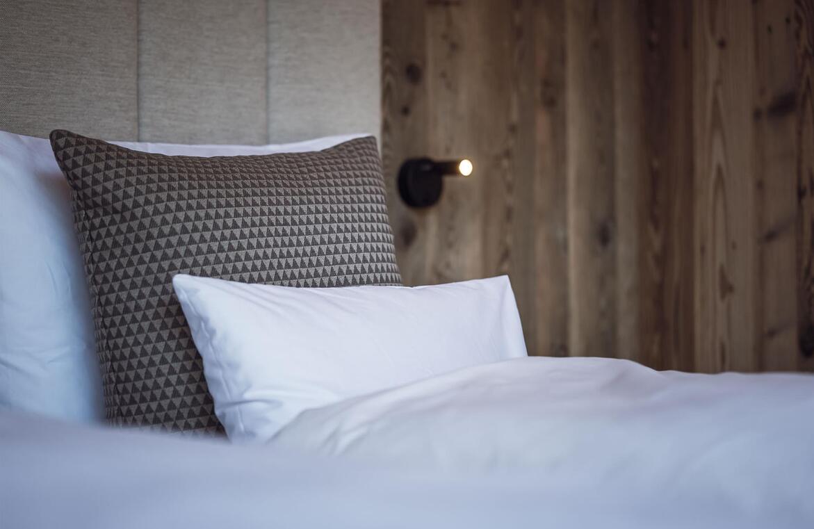 Doppelbett mit weißer Bettwäsche, braun gemusterten Kissen, gepolstertem Kopfteil an einer Holzwand.