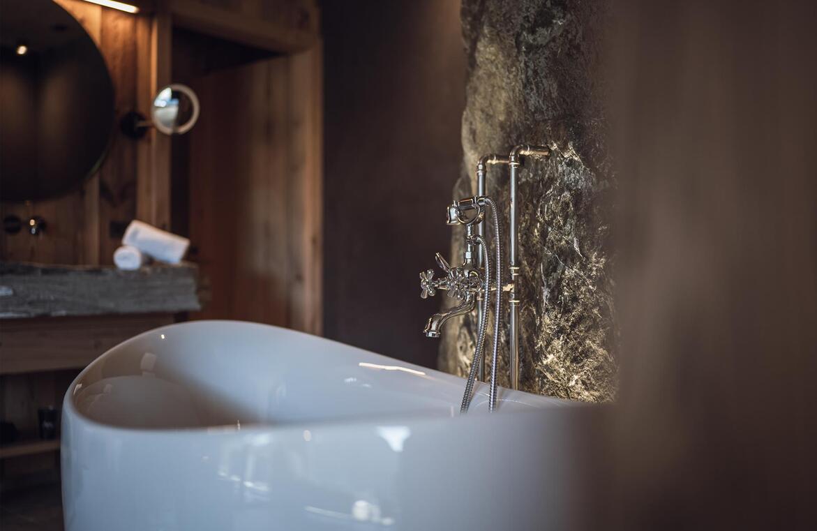 Vasca da bagno freestanding bianca con rubinetteria argentata e doccetta davanti a una parete rocciosa illuminata.