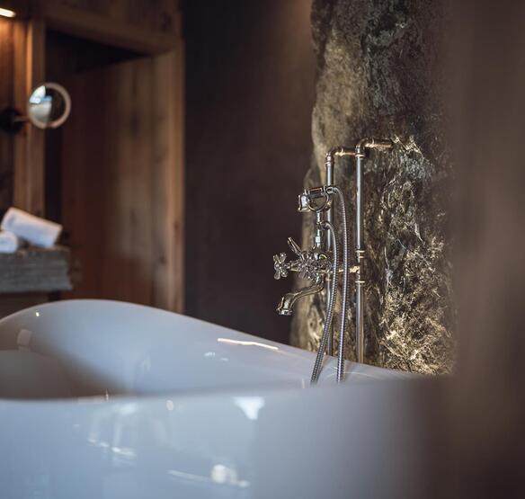 Vasca da bagno freestanding bianca con rubinetteria argentata e doccetta davanti a una parete rocciosa illuminata.
