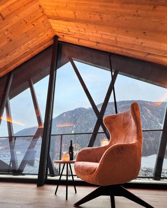 Polstersessel neben einem kleinen runden Beistelltisch vor großer Glasfront mit Blick auf die verschneite Berglandschaft.