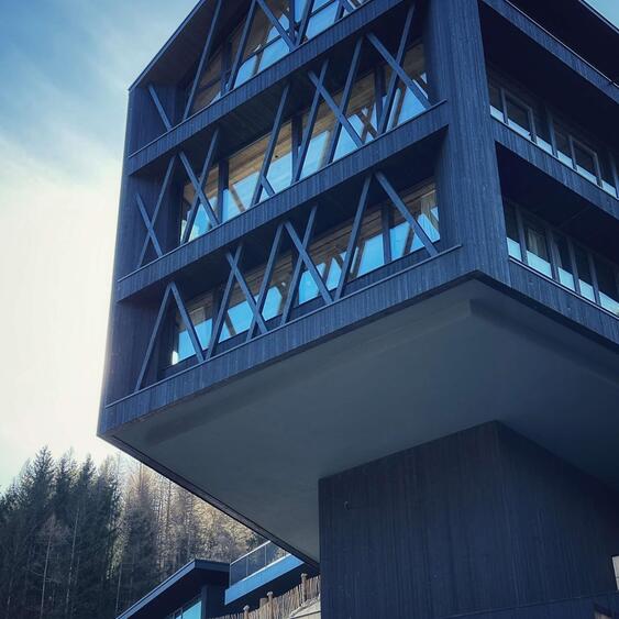 Moderne Fassade der Amus Chalets aus dunklem Holz mit großen Glasfronten und diagonalen Holzelementen vor bewaldetem Hang.