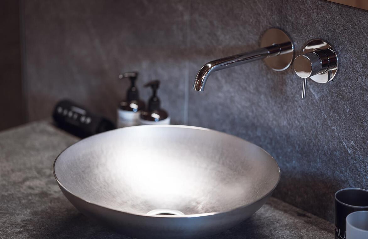 Rundes Aufsatzwaschbecken mit Wandarmatur auf einer grauen Waschtischplatte, daneben Pflegeprodukte.