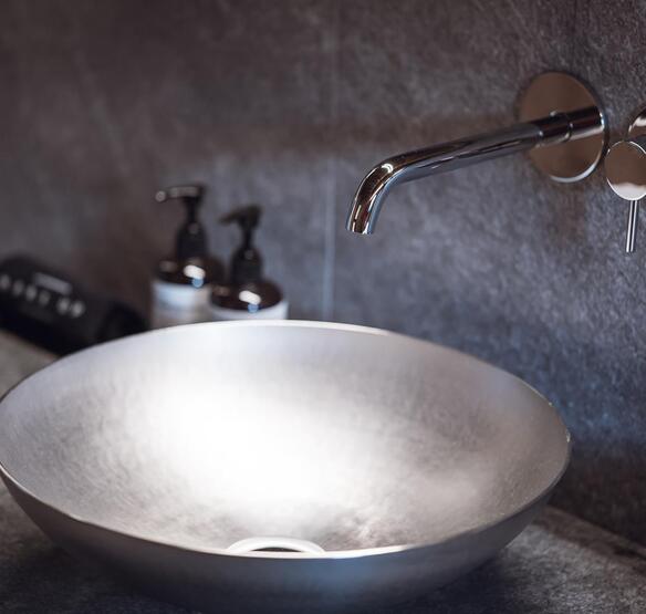 Lavabo rotondo da appoggio con rubinetteria a parete su un piano grigio, con prodotti per la cura del corpo accanto.