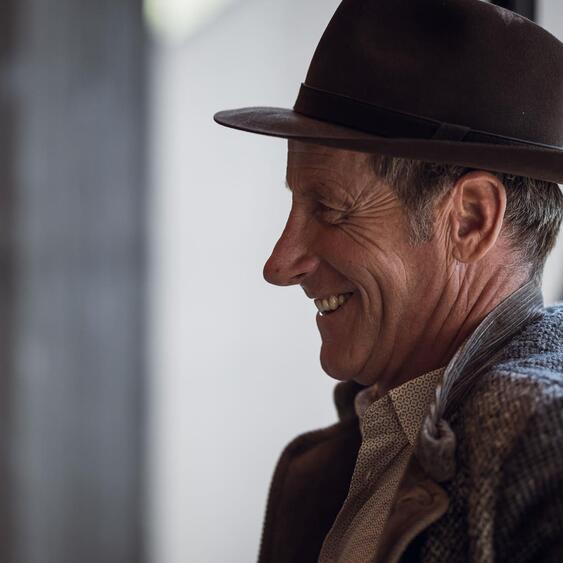 Uomo anziano sorridente, di profilo, con cappello scuro e cardigan grigio.