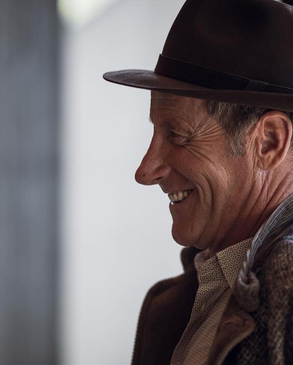 Smiling elderly man in profile, wearing a dark hat and grey cardigan.