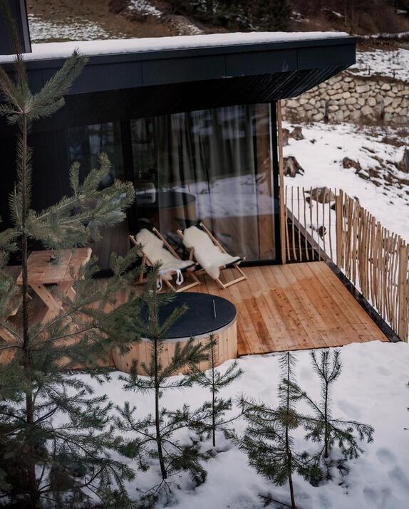 Amus Chalet mit Holzterrasse, Außenwhirlpool und zwei Liegestühlen in verschneiter Landschaft.