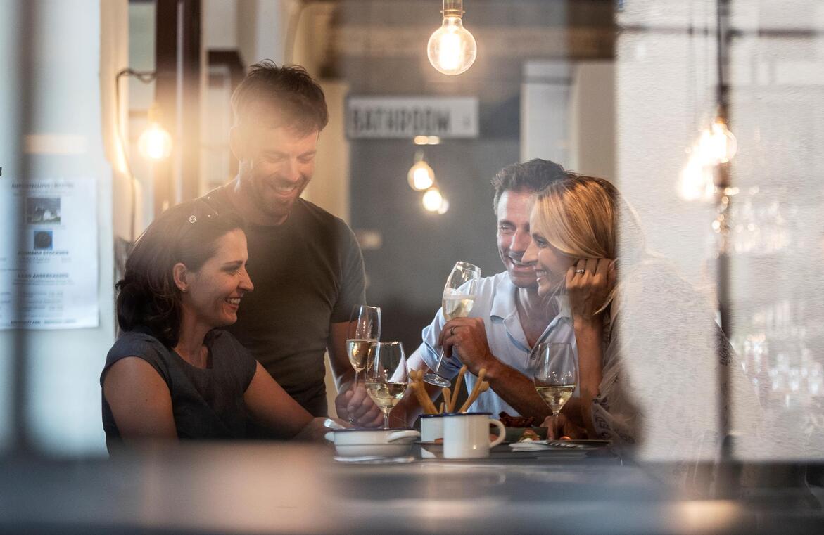 View through a window into a brightly lit room, where several people are sitting at a table and drinking wine.