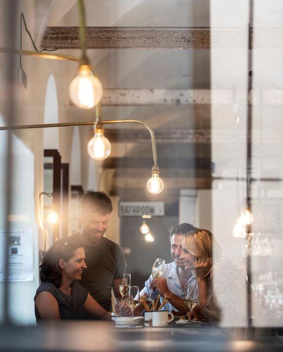 View through a window into a brightly lit room, where several people are sitting at a table and drinking wine.