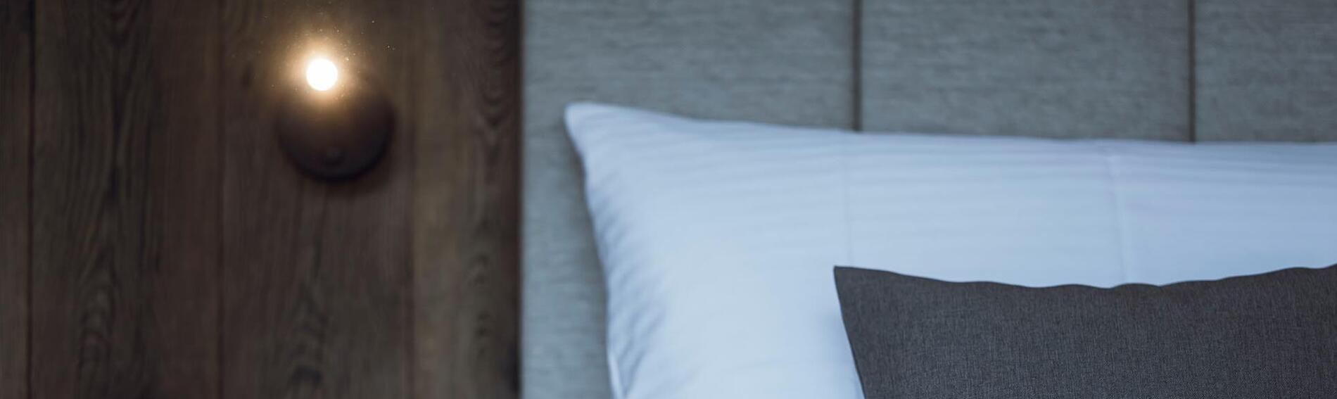 Close-up view of a chalet bed with a white pillow, grey decorative cushion, upholstered headboard and lamp on a dark wooden wall.
