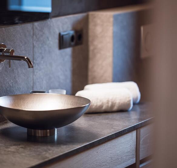 Lavabo rotondo da appoggio con rubinetteria a parete su un piano grigio, con asciugamani bianchi arrotolati accanto.
