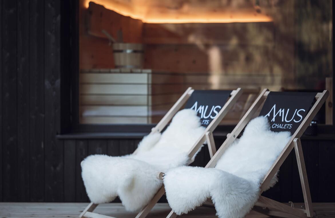 Liegestühle mit Amus Chalet Logo und weißem Fell vor einer Glasscheibe mit Blick in die beleuchtete Sauna.