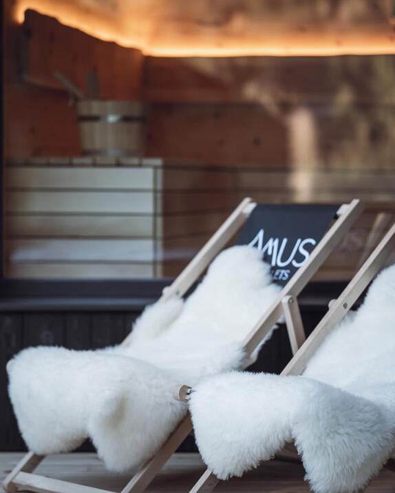 Liegestühle mit Amus Chalet Logo und weißem Fell vor einer Glasscheibe mit Blick in die beleuchtete Sauna.