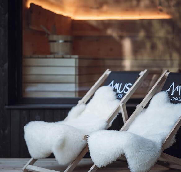 Sedie a sdraio con il logo Amus Chalet e una pelliccia bianca davanti a una vetrata con vista sulla sauna illuminata.