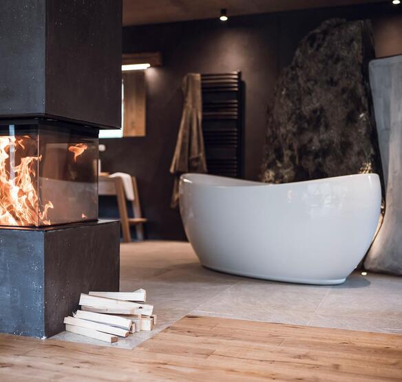 Vasca da bagno freestanding davanti a una parete rocciosa, con camino acceso in primo piano.