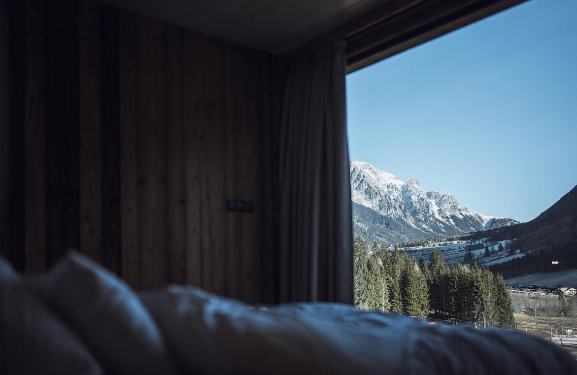 Blick vom Bett durch ein großes Panoramafenster auf schneebedeckte Berge und Wälder.