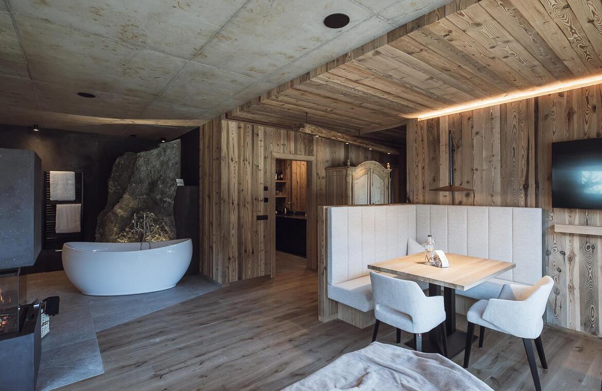 Chalet-Wohnbereich mit Kamin, Holztisch, gepolsterten Sitzmöbeln und freistehender Badewanne im Hintergrund.