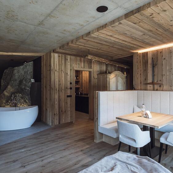 Chalet-Wohnbereich mit Kamin, Holztisch, gepolsterten Sitzmöbeln und freistehender Badewanne im Hintergrund.