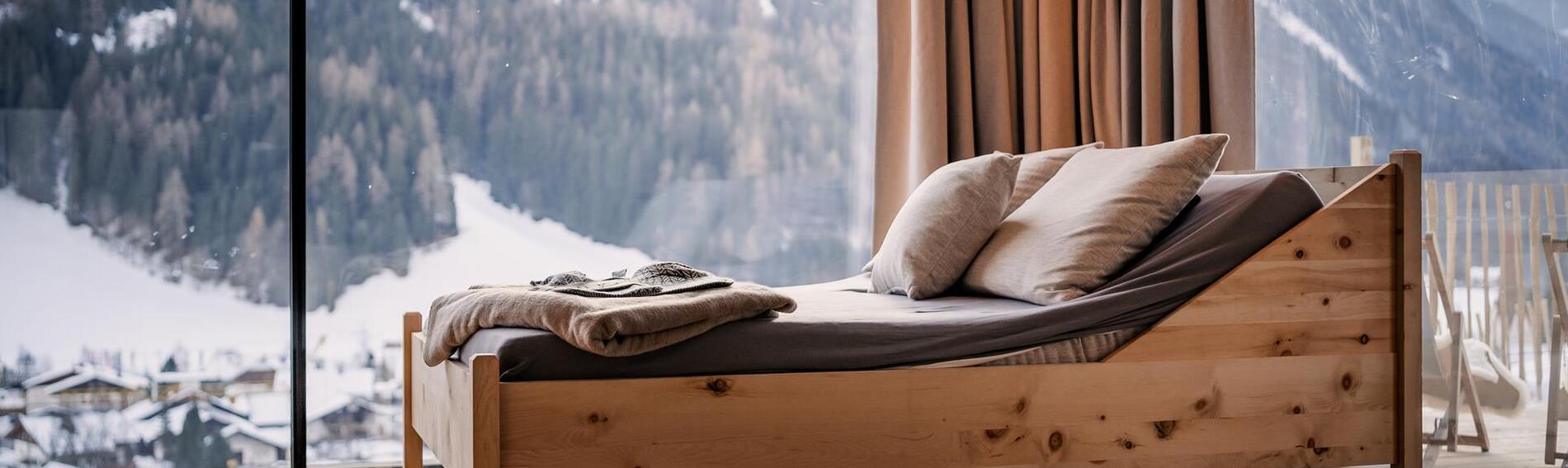 Lettino in legno con cuscino e coperta davanti a un'ampia vetrata con vista sul paesaggio montano innevato.