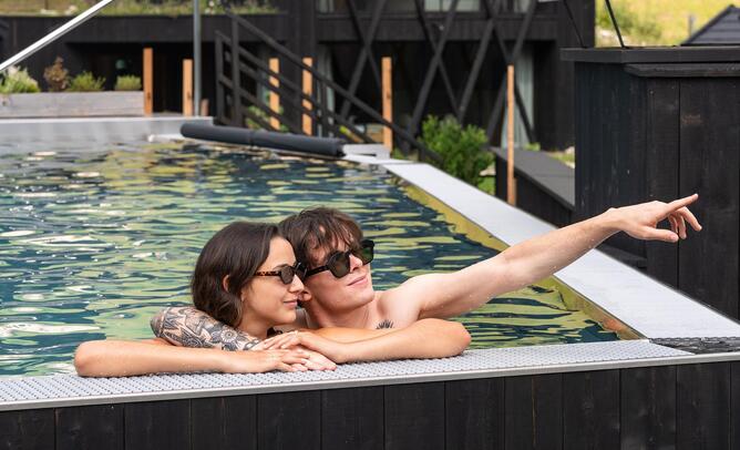 Zwei Personen relaxen im Außenpools mit dunklen Holzchalets und einer grünen Wiese im Hintergrund.