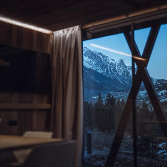Essbereich im Chalet mit großer Fensterfront und Blick auf schneebedeckte Berge in der Dämmerung.