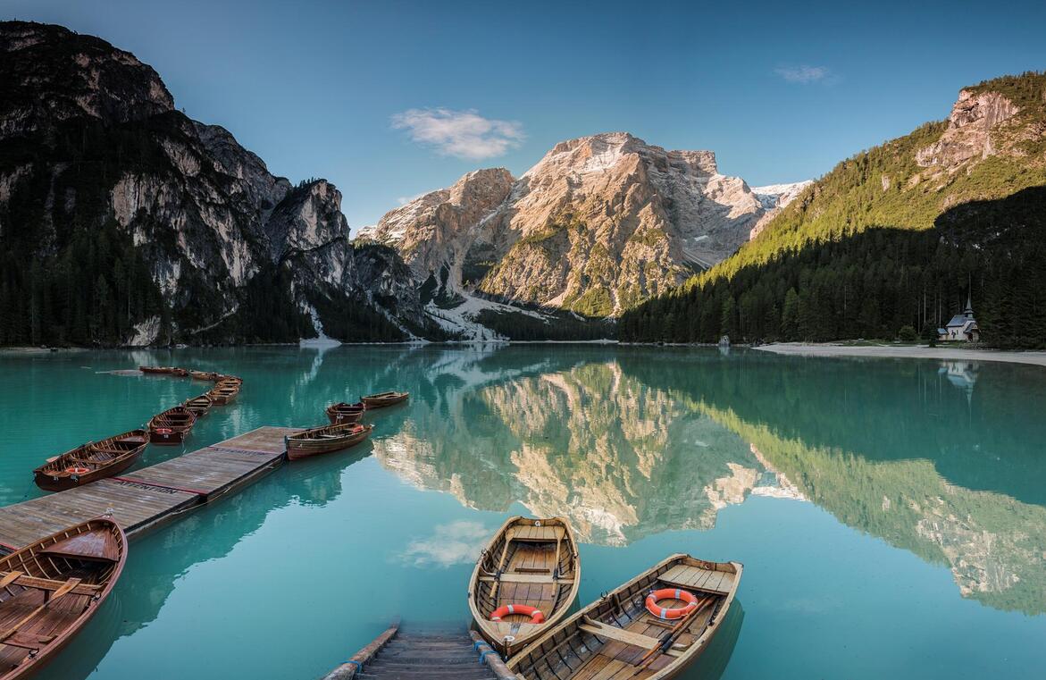Lago di Braies al sole con barche e una cappella, boschi e montagne sullo sfondo.