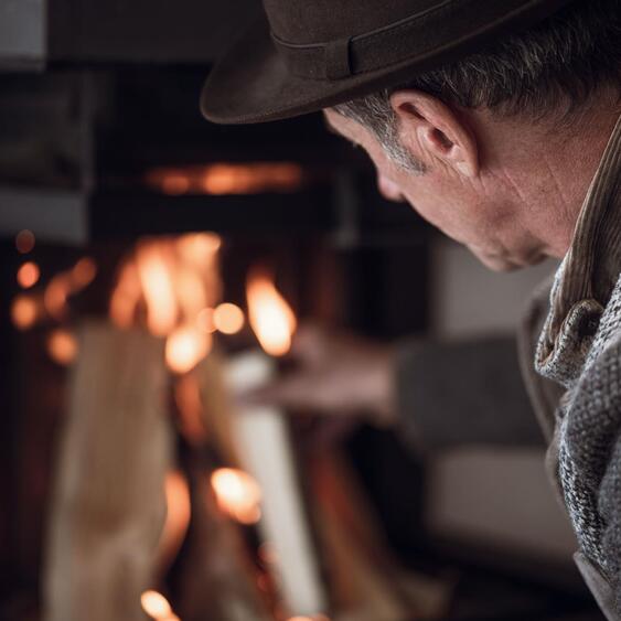 Älterer Mann mit dunklem Hut und grauer Strickjacke legt Holz in den brennenden Kamin.