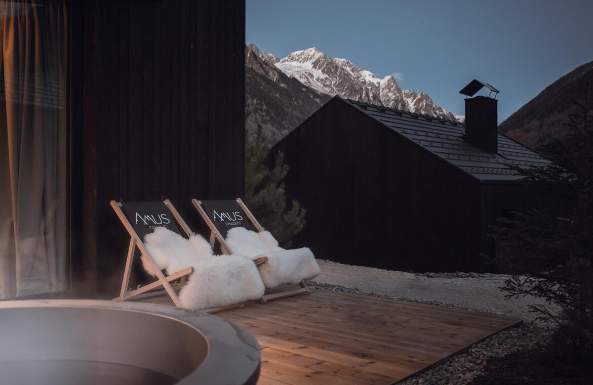 Zwei Liegestühle mit Amus Chalets Logo und weißem Fell auf einer Holzterrasse mit Blick auf die schneebedeckten Berge in der Dämmerung.