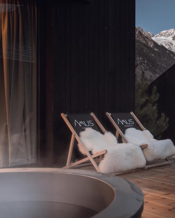 Zwei Liegestühle mit Amus Chalets Logo und weißem Fell auf einer Holzterrasse mit Blick auf die schneebedeckten Berge in der Dämmerung.