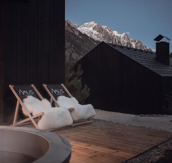 Due sedie a sdraio con il logo Amus Chalets e pellicce bianche su una terrazza in legno con vista sulle montagne innevate al crepuscolo.
