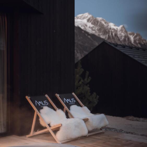 Two deckchairs with the Amus Chalets logo and white fur throws on a wooden terrace overlooking snow-covered mountains at dusk.