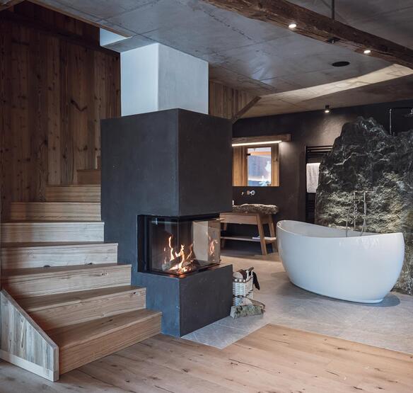 Chalet con scala in legno, camino acceso e vasca da bagno freestanding davanti a una parete rocciosa.