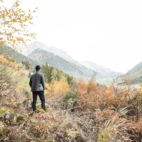 Mann mit Hut steht in einem herbstlichen Wald mit Blick in die Berge.