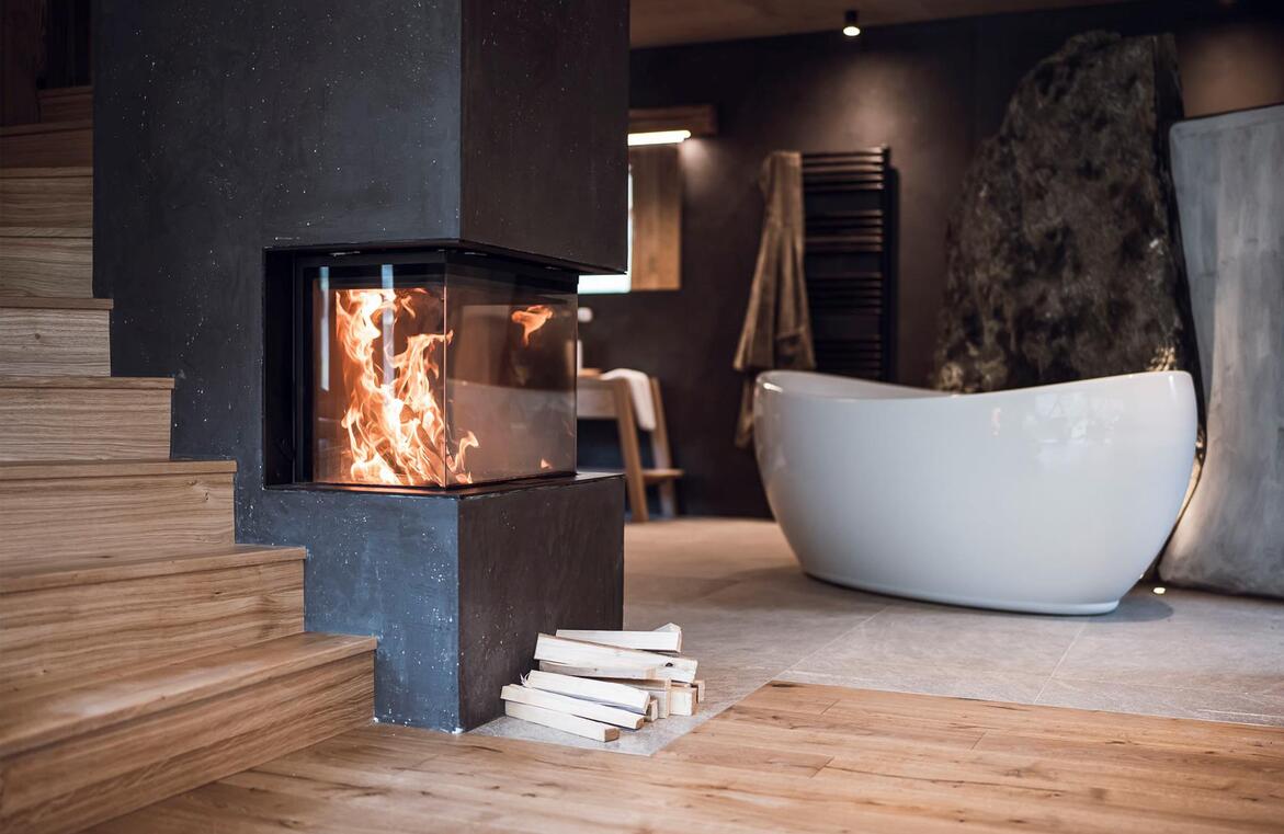 Freistehende Badewanne an einer Felswand mit brennendem Kamin im Vordergrund.