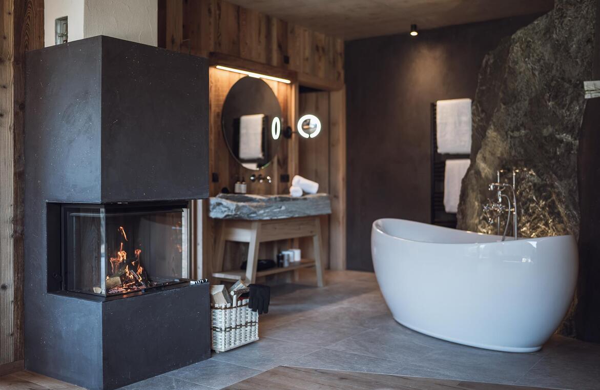 Vasca da bagno freestanding davanti a una parete rocciosa, lavabo in pietra naturale e camino acceso.