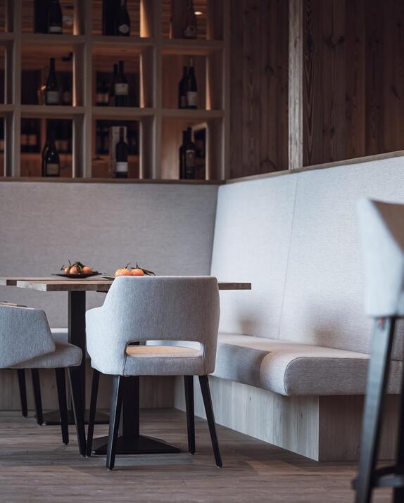 Bar- und Essbereich mit hell gepolsterten Sitzmöbeln, Holztischen und Blick auf das offene Weinregal.