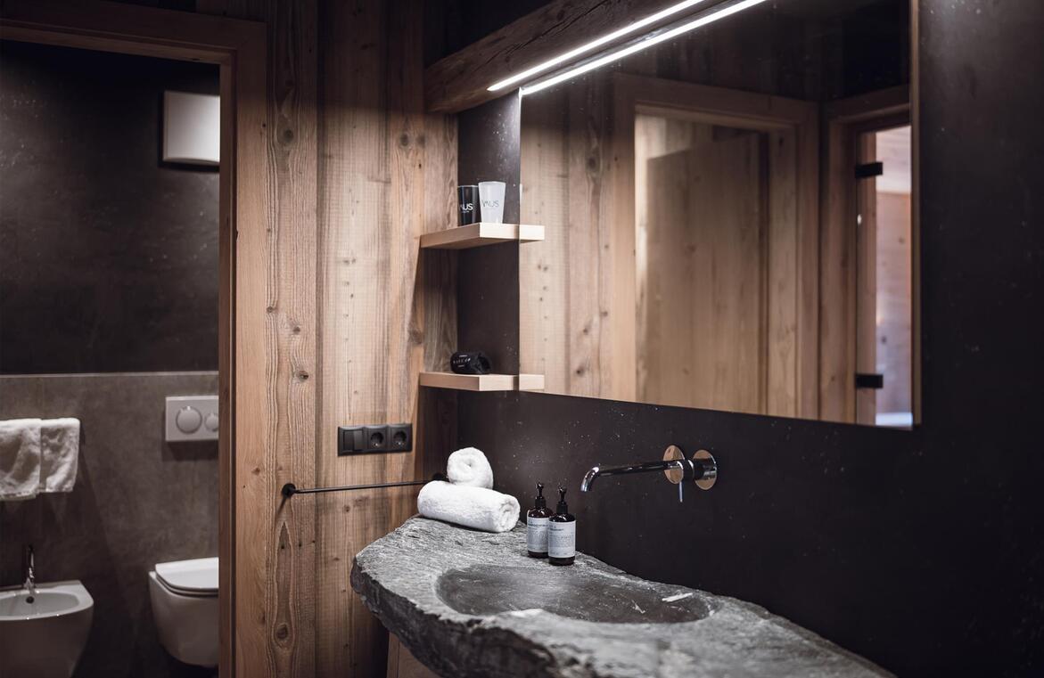 Bagno dello chalet con lavabo in pietra, grande specchio, WC, bidet, piastrelle nere e grigie ed elementi in legno.