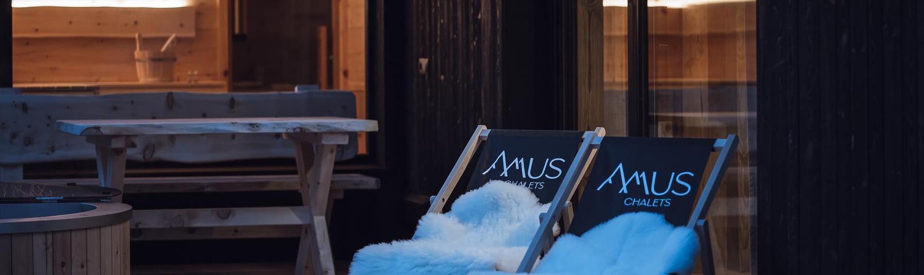 Sedie a sdraio con il logo Amus Chalet e una pelliccia bianca davanti a una vetrata con vista sulla sauna illuminata.