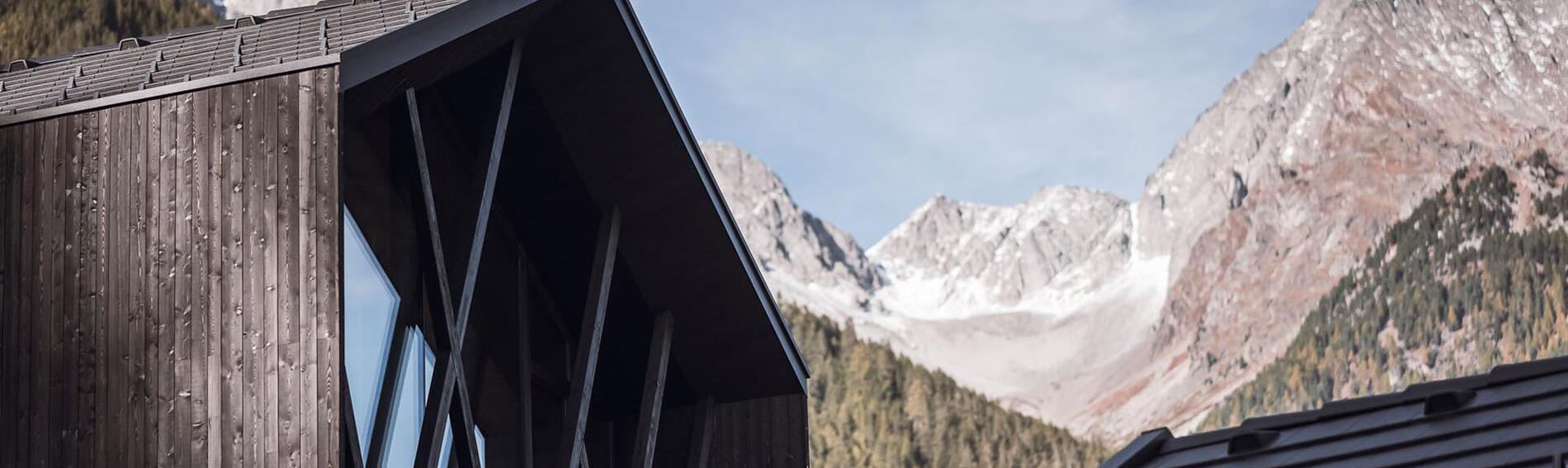Dunkle Holzfassade eines Chalets mit großen Fenstern und Holzbalken, im Hintergrund massive Berglandschaft.