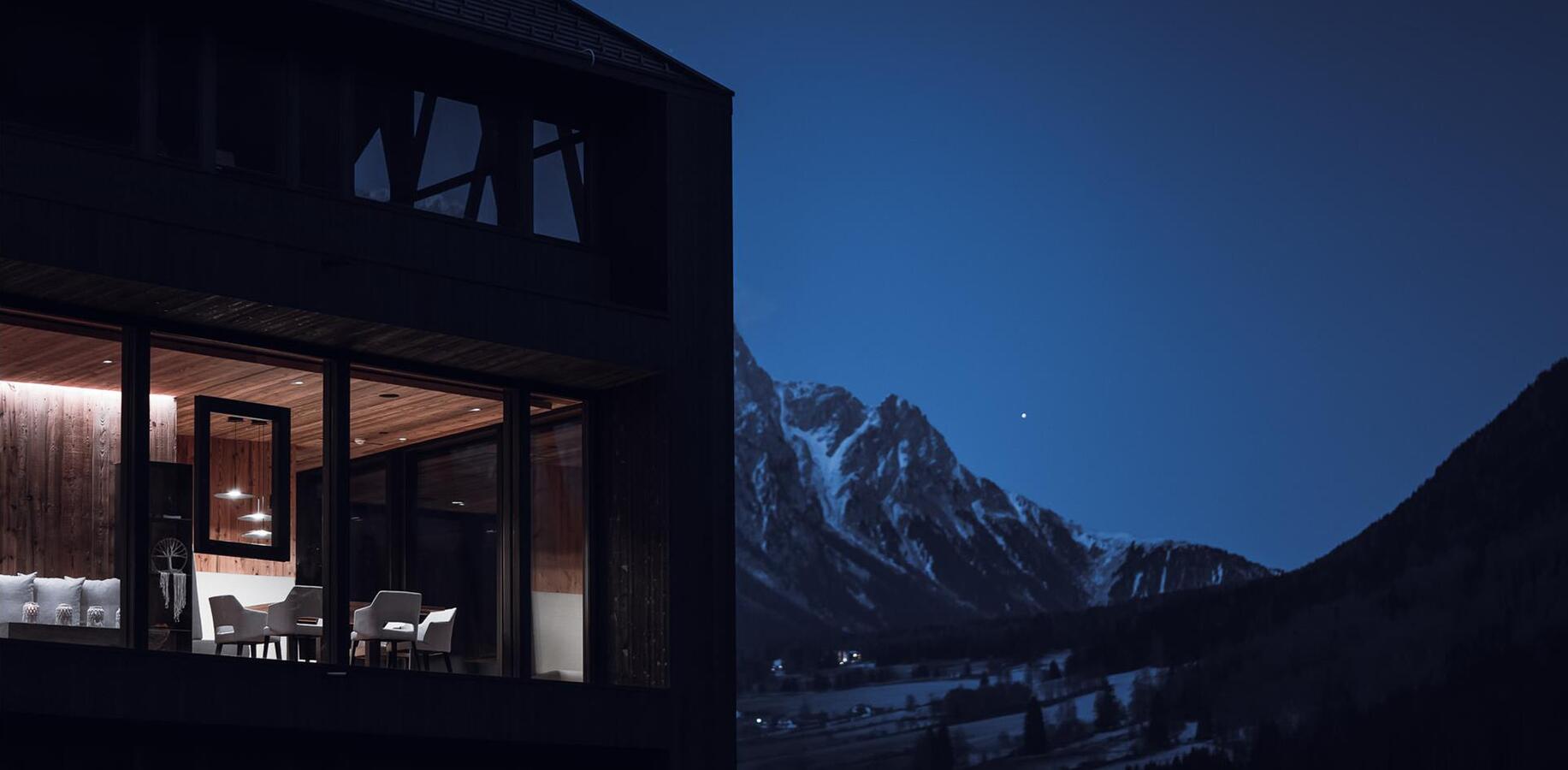 Dark wooden chalet with an illuminated glass façade and seating area, with snow-covered mountains in the background at dusk.