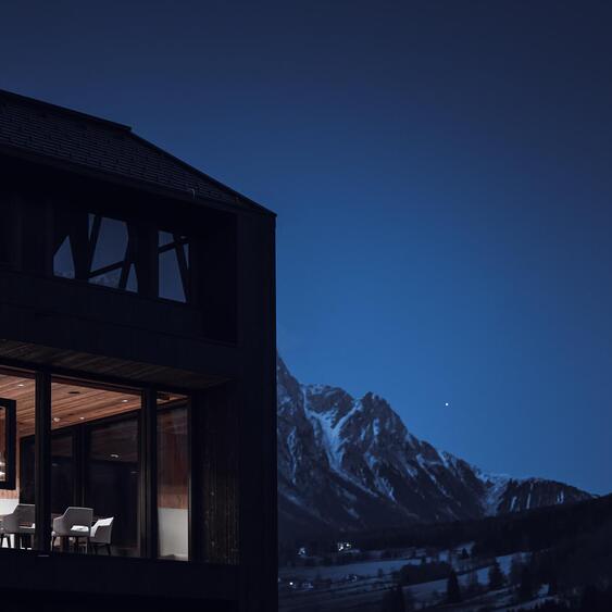 Dunkles Holzchalet mit beleuchteter Glasfront und Sitzbereich, im Hintergrund schneebedeckte Berge in der Abenddämmerung.