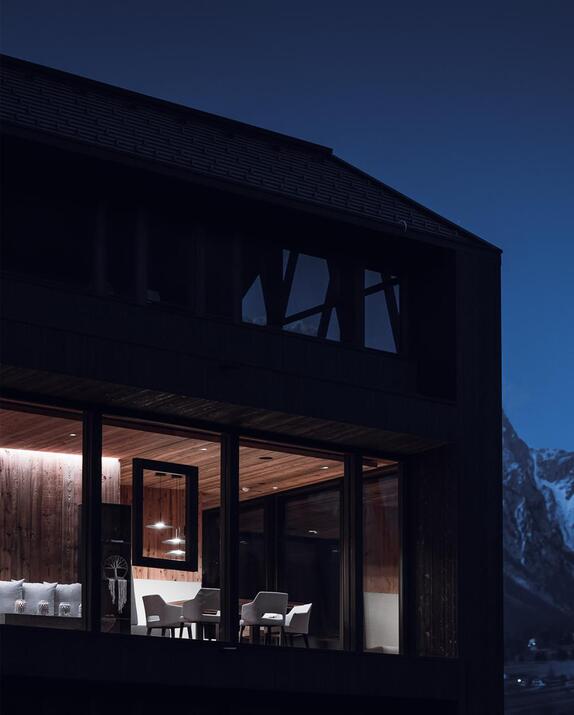 Dunkles Holzchalet mit beleuchteter Glasfront und Sitzbereich, im Hintergrund schneebedeckte Berge in der Abenddämmerung.