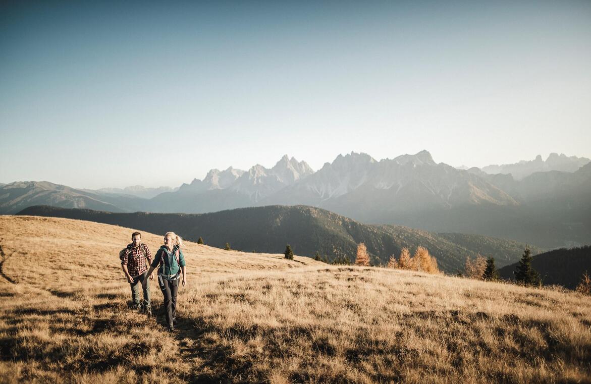 Due escursionisti su un sentiero tra prati alpini autunnali, con un imponente paesaggio montano sullo sfondo.