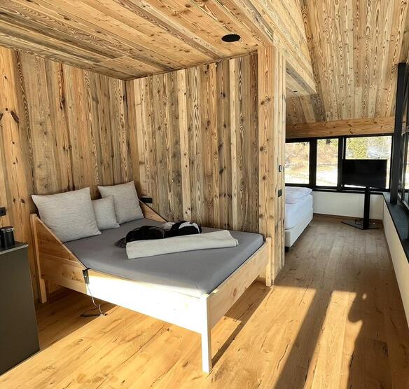 Zona giorno e notte degli Amus Chalets con rivestimenti in legno, divano, letto matrimoniale e ampie finestre panoramiche.