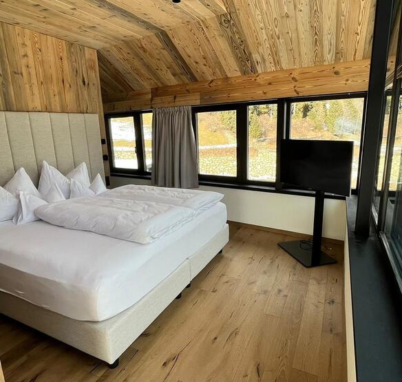 Camera da letto degli Amus Chalets con letto matrimoniale, rivestimenti in legno chiaro, televisore e ampie finestre.