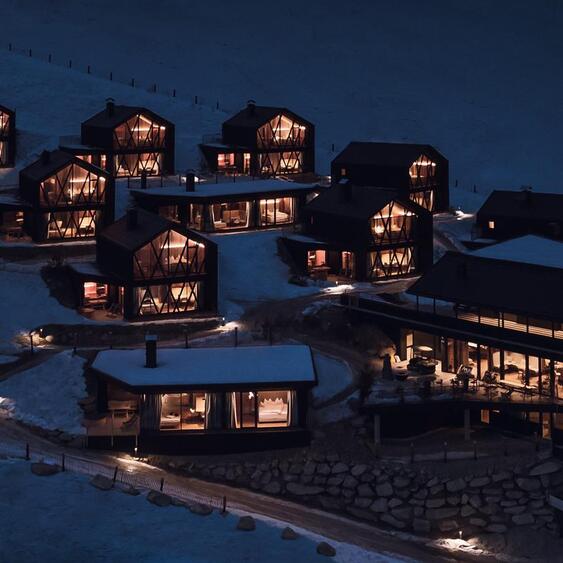 Vista aerea degli Amus Chalets illuminati su un pendio montano innevato al crepuscolo.