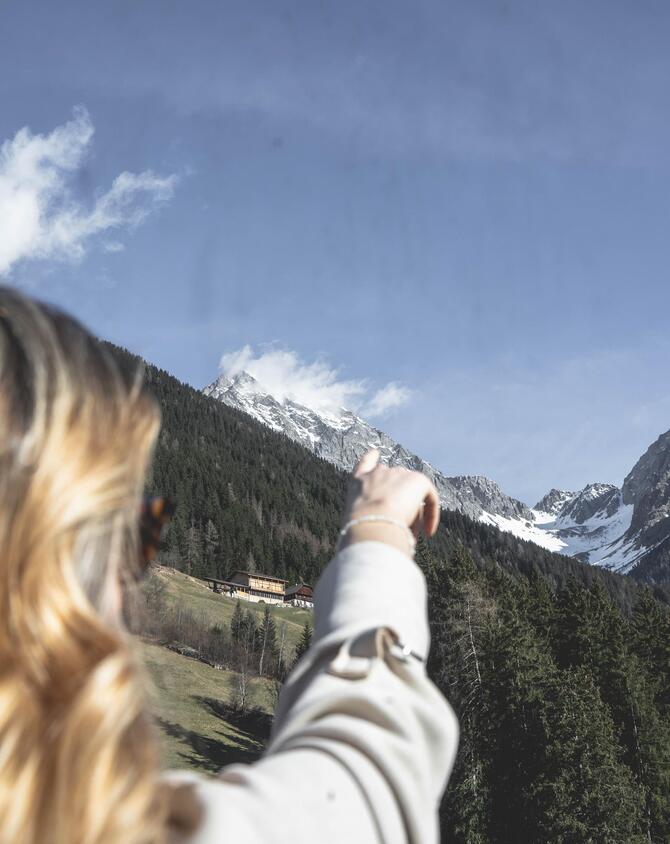 Frau zeigt auf schneebedeckte Berggipfel mit bewaldeten Hängen unter blauem Himmel.