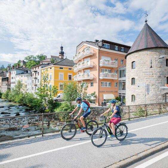 Zwei Mountainbiker fahren entlang eines Flusses durch eine Stadt mit historischen Gebäuden und einem steinernen Turm.