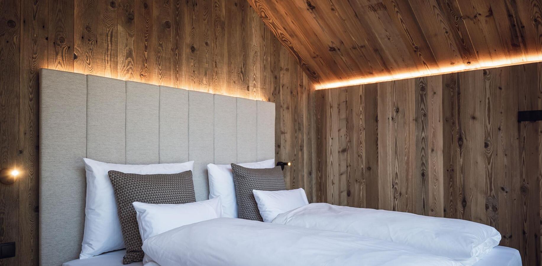 Doppelbett mit weißer Bettwäsche, gepolstertem Kopfteil in einem holzverkleideten Schlafzimmer mit indirekter Beleuchtung.