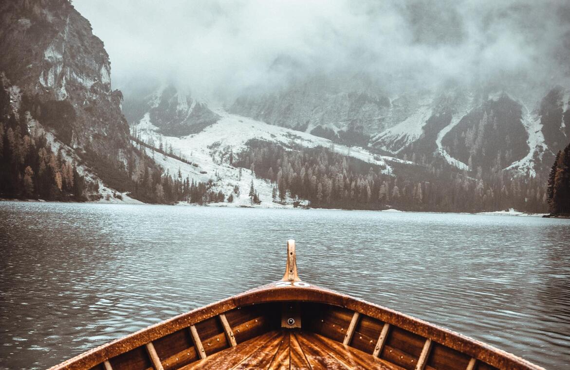 Blick vom Bug eines Holzbootes auf einen Bergsee, umgeben von bewaldeter und schneebedeckter Landschaft im Nebel.