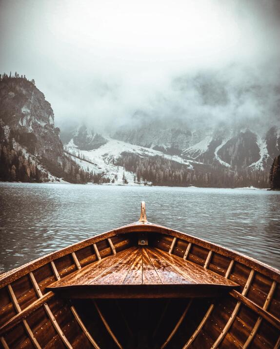 View from the bow of a wooden boat overlooking a mountain lake, surrounded by a misty landscape of forests and snow.